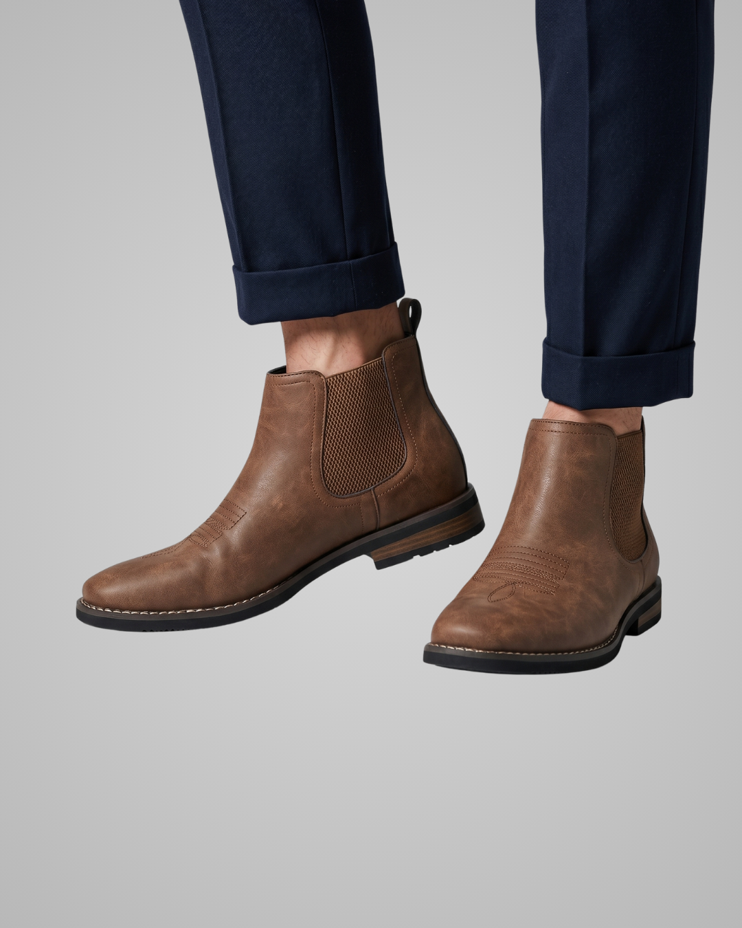 Brown leather Chelsea boots worn with navy pants on a light gray background