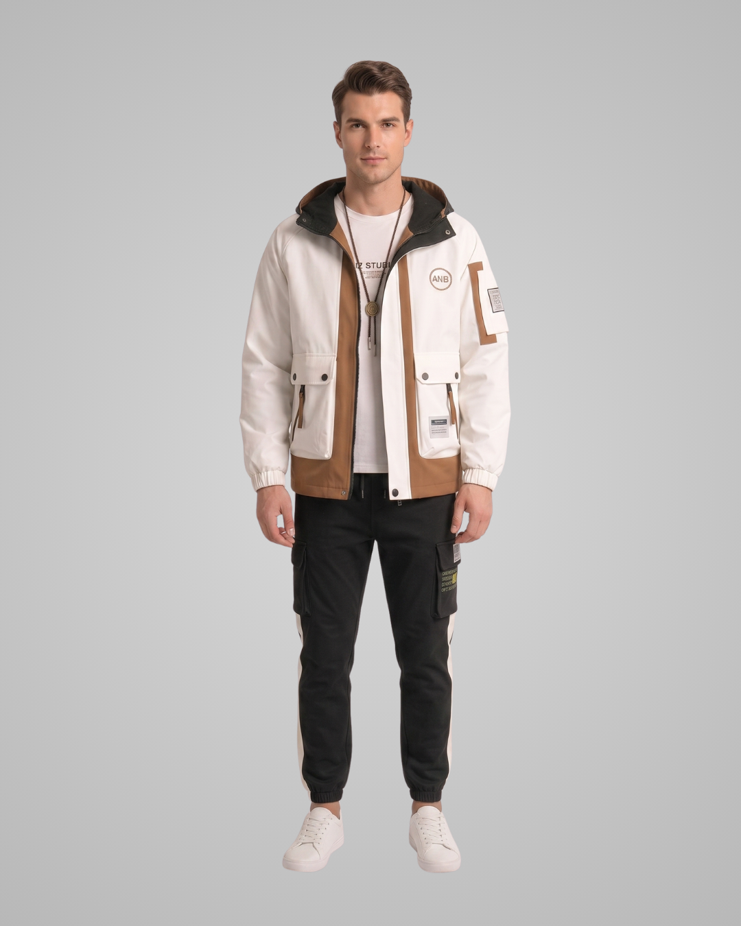 A man modeling a beige and brown technical hooded jacket with a white t-shirt, black cargo pants, and white sneakers, set against a solid gray studio background.