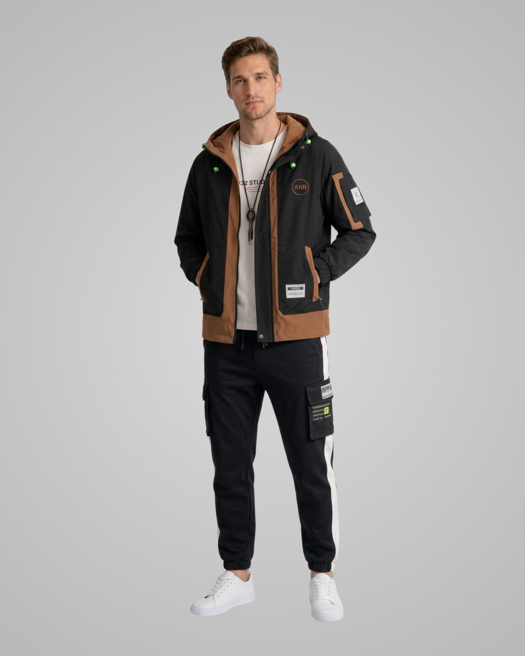 A man modeling a black and brown hooded jacket with green accents, black cargo pants with white side stripes, and white sneakers, set against a solid gray background.