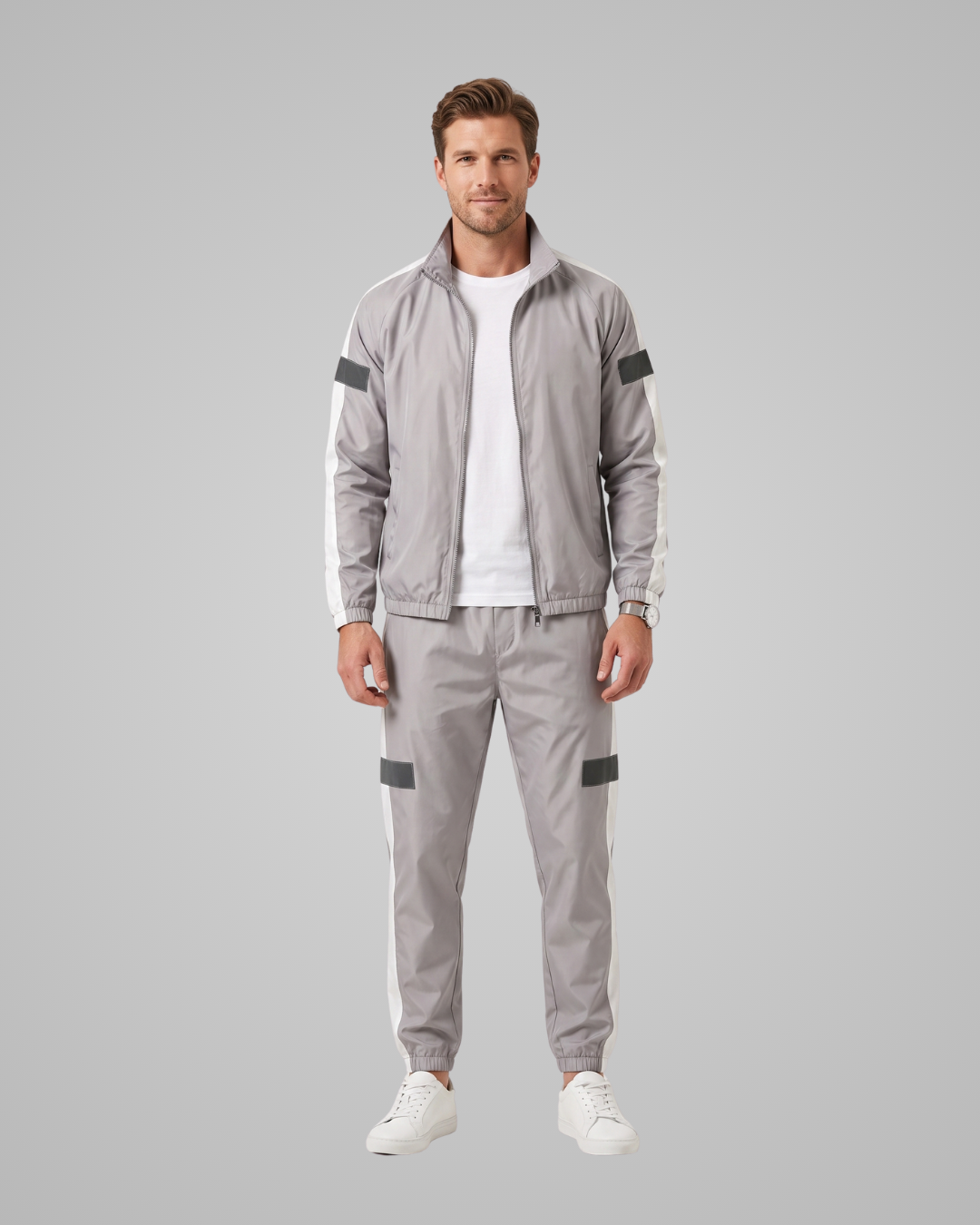 A man modeling a light gray athletic tracksuit with white side stripes and dark gray accent panels, worn over a white t-shirt and paired with white sneakers, against a solid gray studio background.