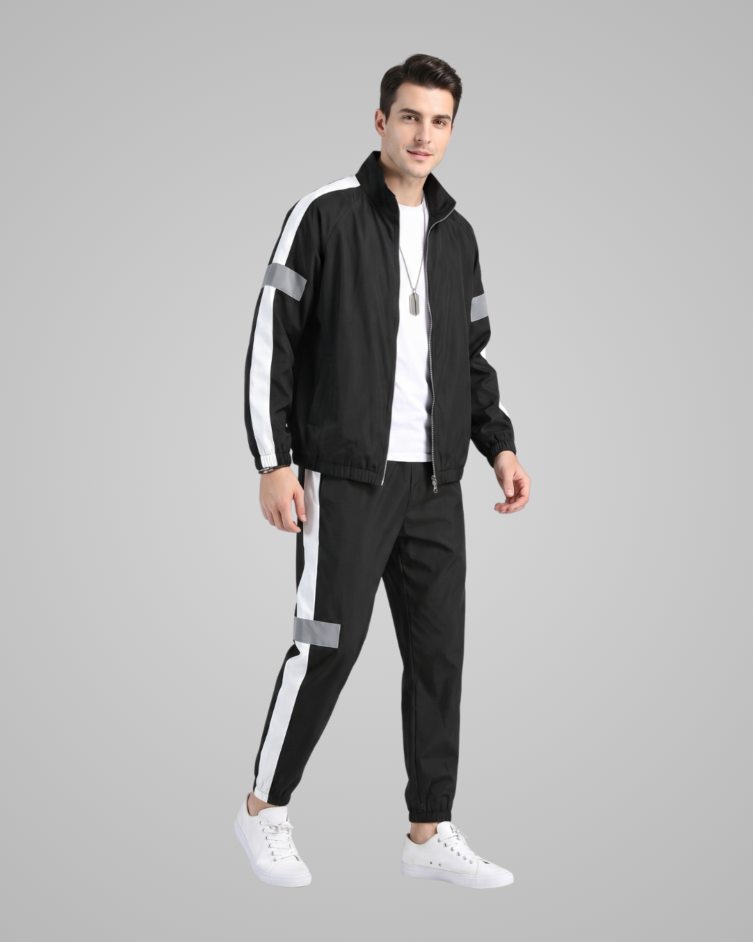 A man modeling a black tracksuit with white side stripes and reflective gray accents, paired with a white t-shirt and white sneakers, against a solid gray background.