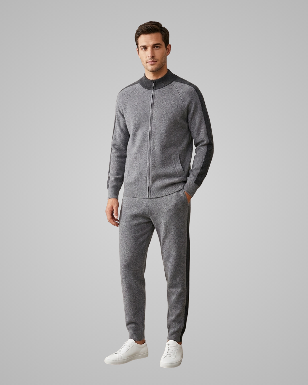 A man modeling a gray heathered zip-up track jacket and matching jogger pants with dark side panels, paired with white sneakers, against a solid gray studio background.