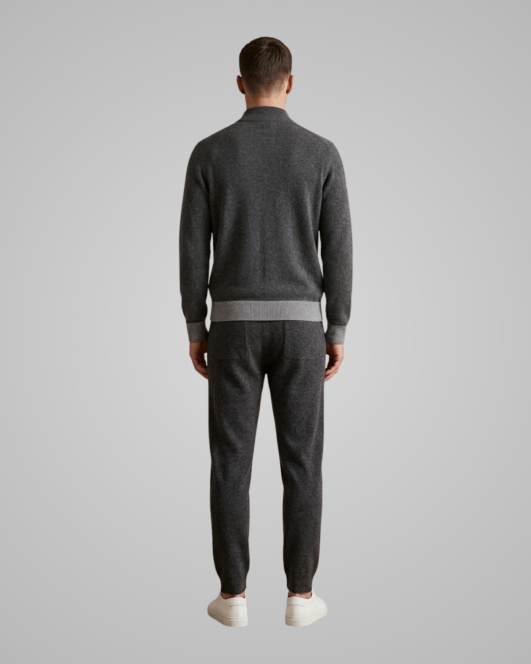 A man viewed from the back wearing a dark gray knit sweater and matching dark gray jogger pants with white sneakers, set against a solid light gray studio background.