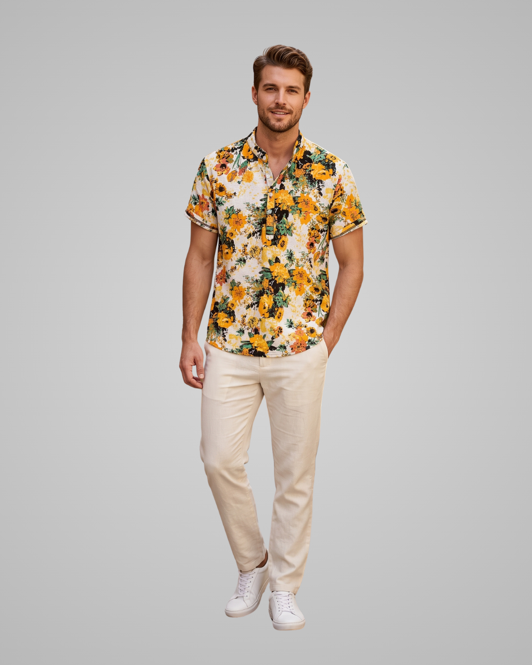 A man modeling a short-sleeve button-up shirt with a vibrant yellow, orange, and green floral print on a cream background, paired with beige trousers and white sneakers, set against a solid gray studio background.