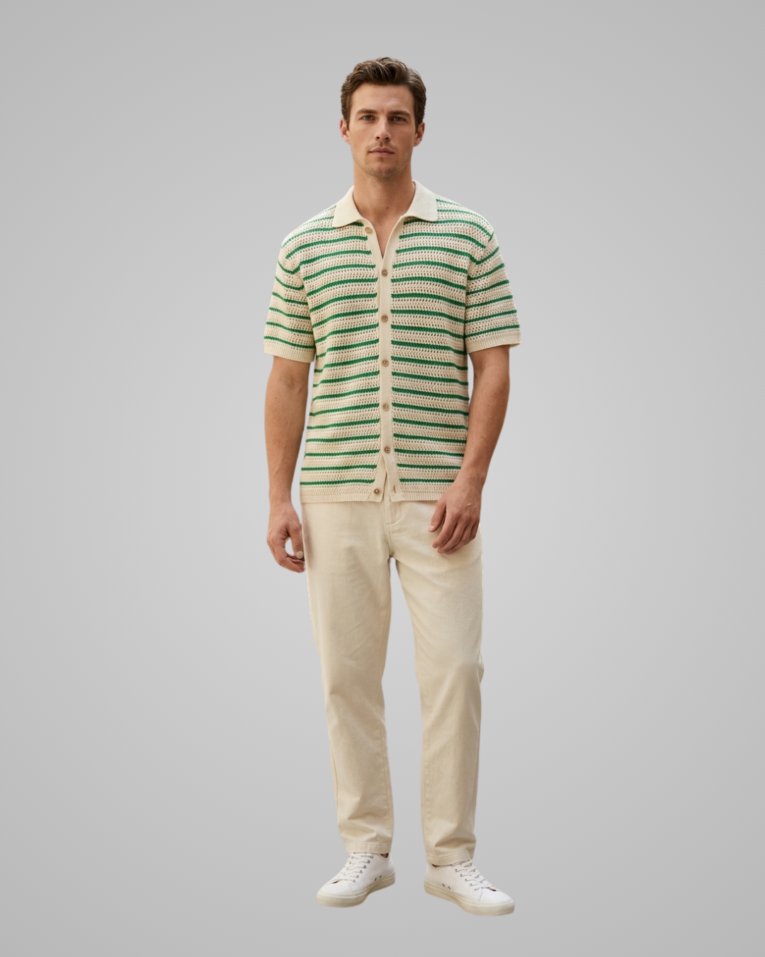 A man modeling a short-sleeve knit polo shirt with green and cream horizontal stripes, paired with cream trousers and white sneakers, against a solid gray studio background.