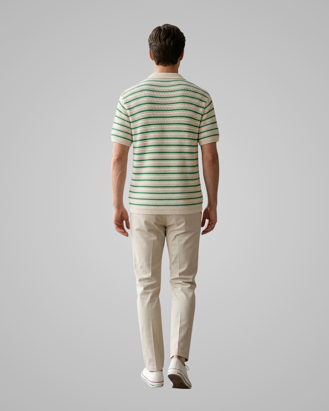 A man viewed from behind wearing a short-sleeve knit polo shirt with thin green and cream horizontal stripes, paired with beige trousers and white sneakers, against a solid light gray studio background.