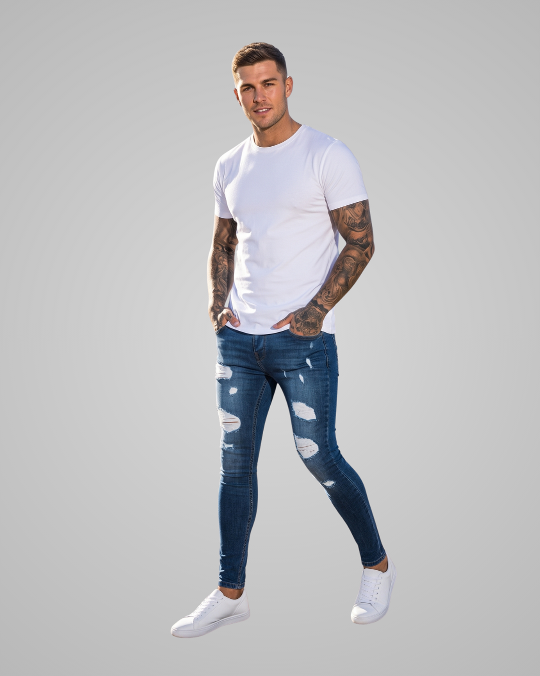 A man modeling a white crew-neck t-shirt and distressed blue skinny jeans with ripped details on the thighs, paired with white sneakers, against a solid gray background.