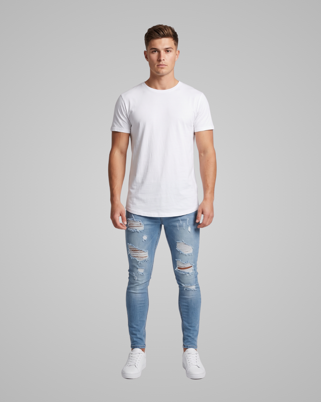 A man modeling a white crew-neck t-shirt and distressed light blue skinny jeans with white sneakers, standing against a solid gray background.