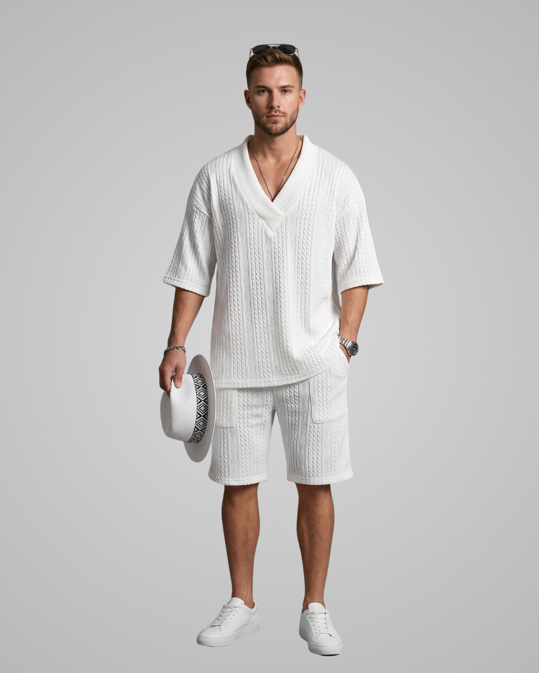 Man wearing a white outfit with shorts and a shirt, holding a hat against a gray background