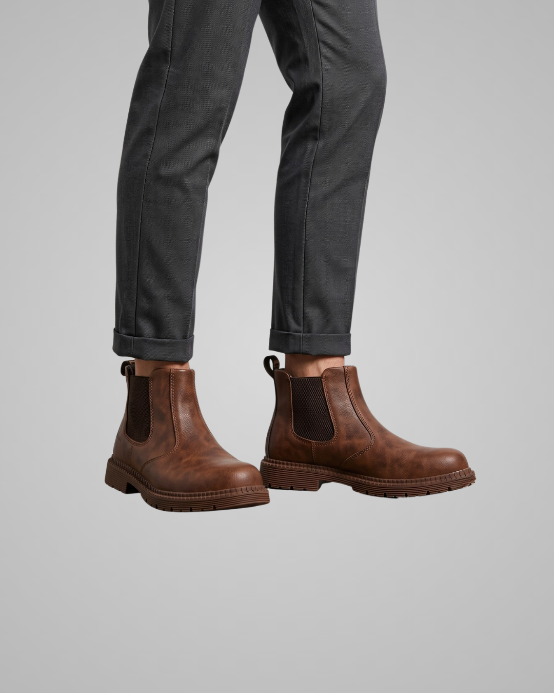 A person wearing brown leather Chelsea-style ankle boots with dark elastic side panels, shown from the calves down with dark gray cuffed pants against a light gray studio background.