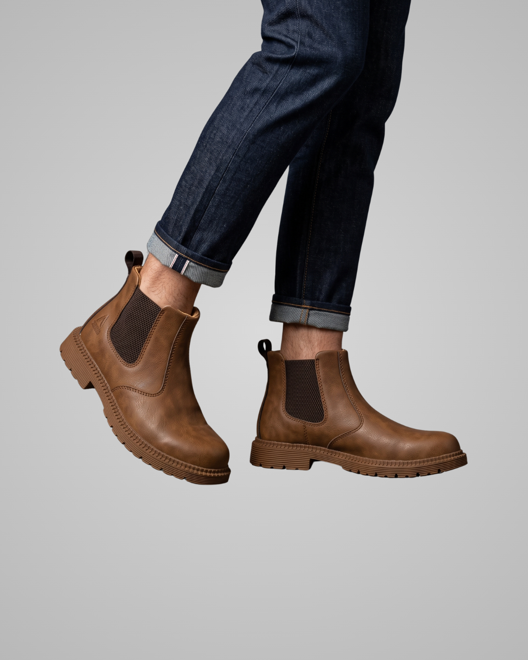 A pair of men's brown leather Chelsea boots worn with dark blue cuffed jeans against a solid gray background.