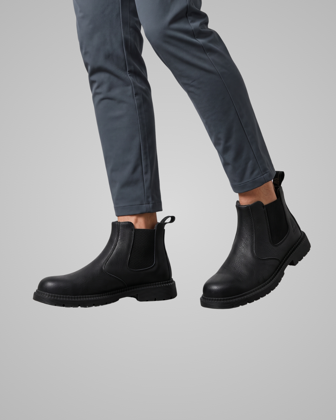 Black leather Chelsea boots worn with dark gray trousers, shown from a side profile against a solid gray background.