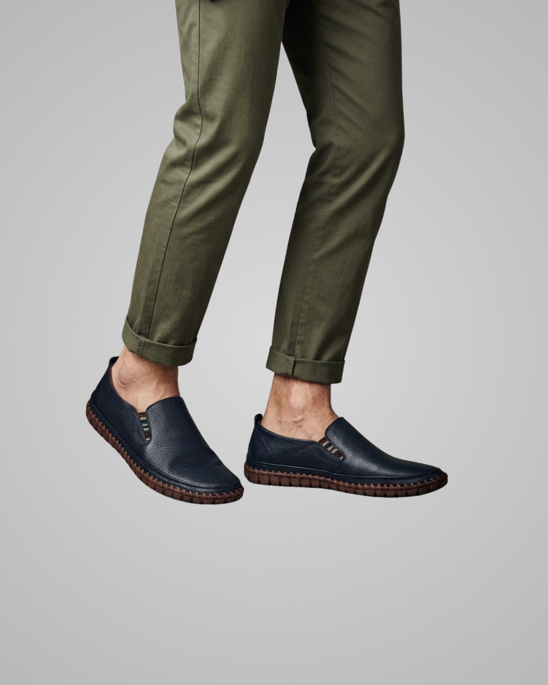 A person wearing olive green cuffed trousers and dark navy slip-on loafers with brown braided soles, shown against a plain light gray studio background.