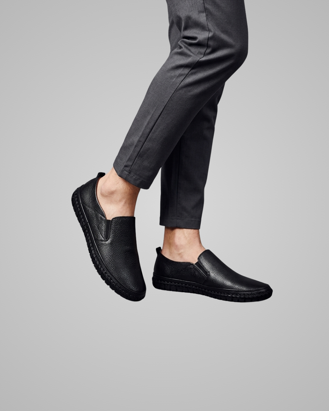 A person wearing black slip-on leather shoes with dark charcoal gray trousers, shown from the calves down against a plain light gray studio background.
