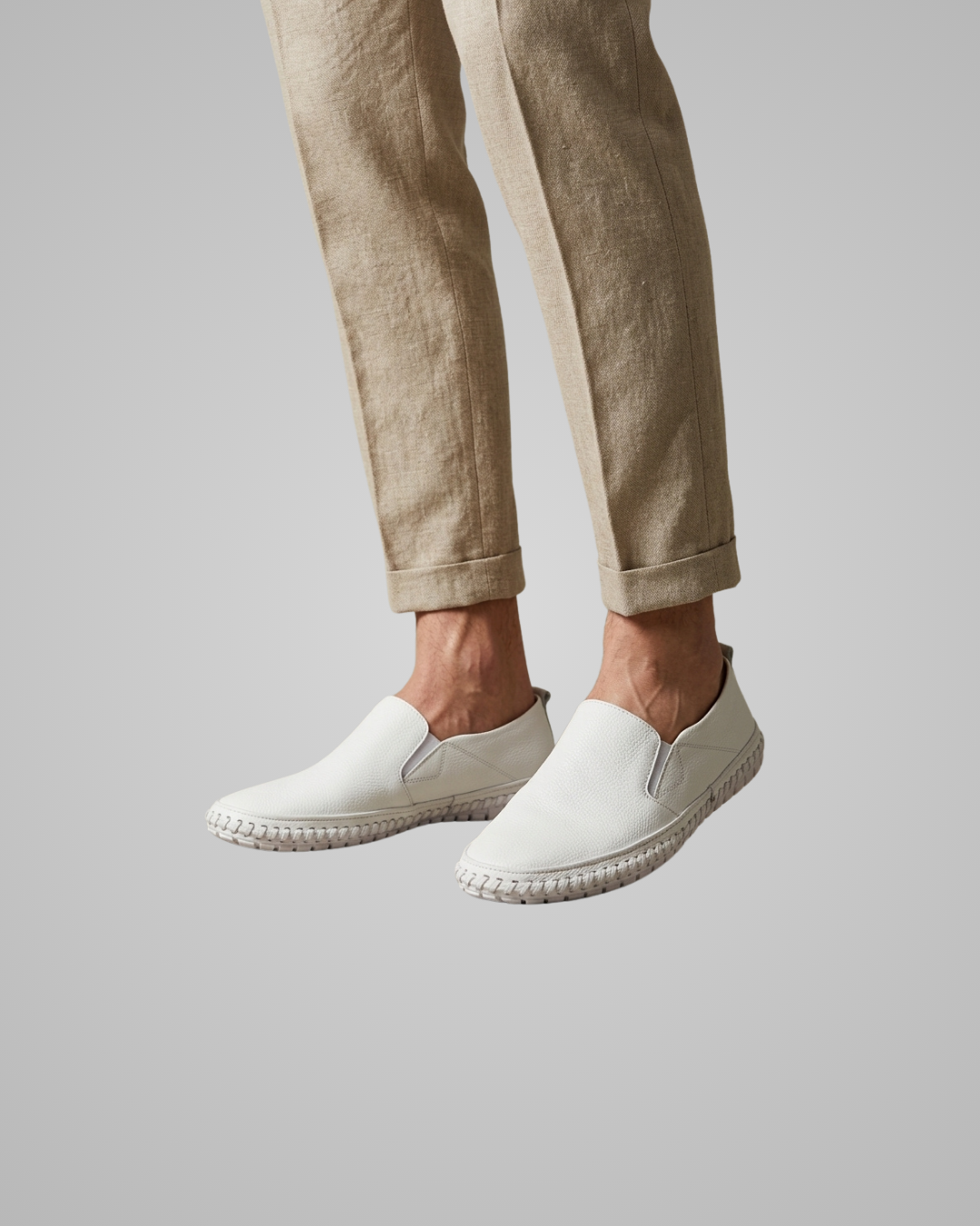 White slip-on espadrille-style shoes worn with light beige trousers, shown against a plain light gray studio background.