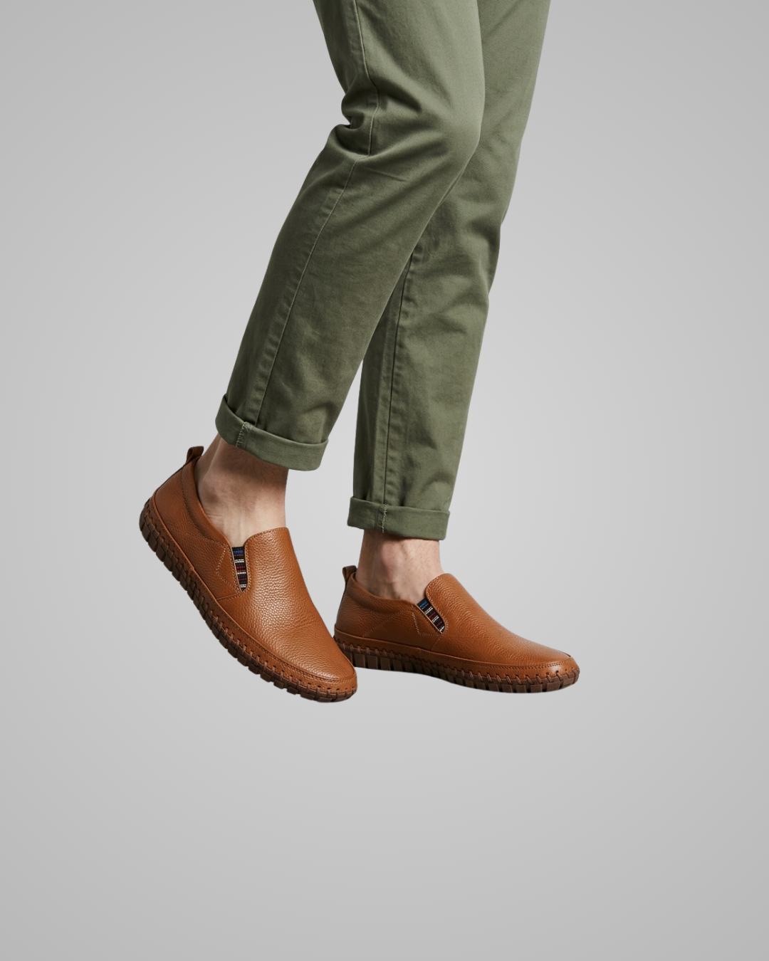 A person wearing olive green pants and brown leather slip-on shoes with small black hardware details, shown against a plain light gray background.