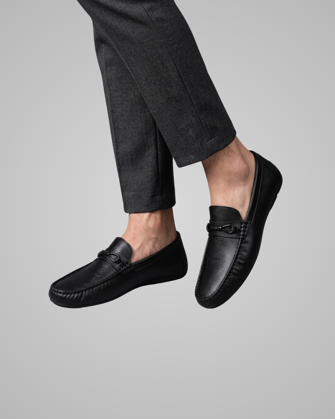 A pair of black leather penny loafers worn with dark charcoal gray trousers, shown against a solid light gray background.