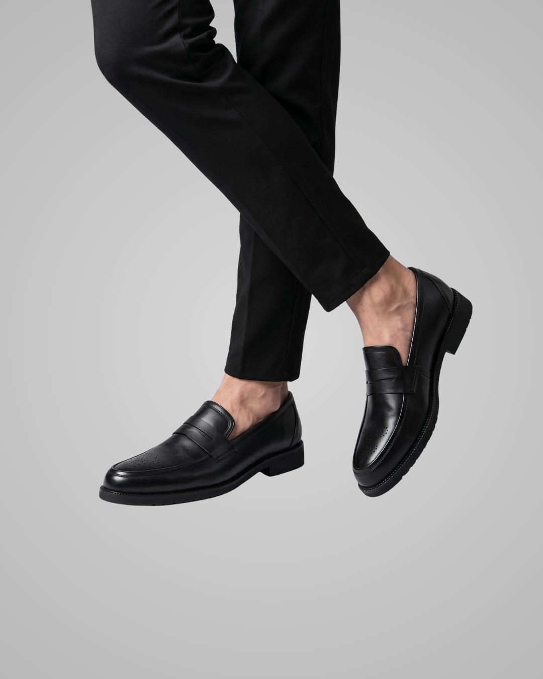 Black leather penny loafers worn with black trousers, shown on a model's feet against a solid gray background.