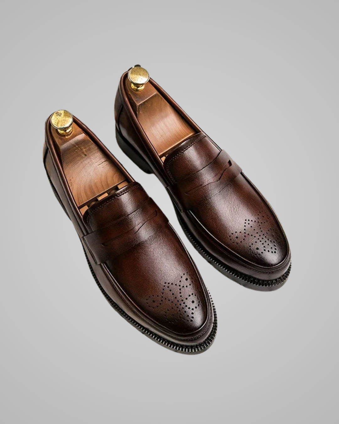 A pair of dark brown leather penny loafers with decorative brogue perforations on the toe, displayed on a light gray background with gold shoe trees inside.