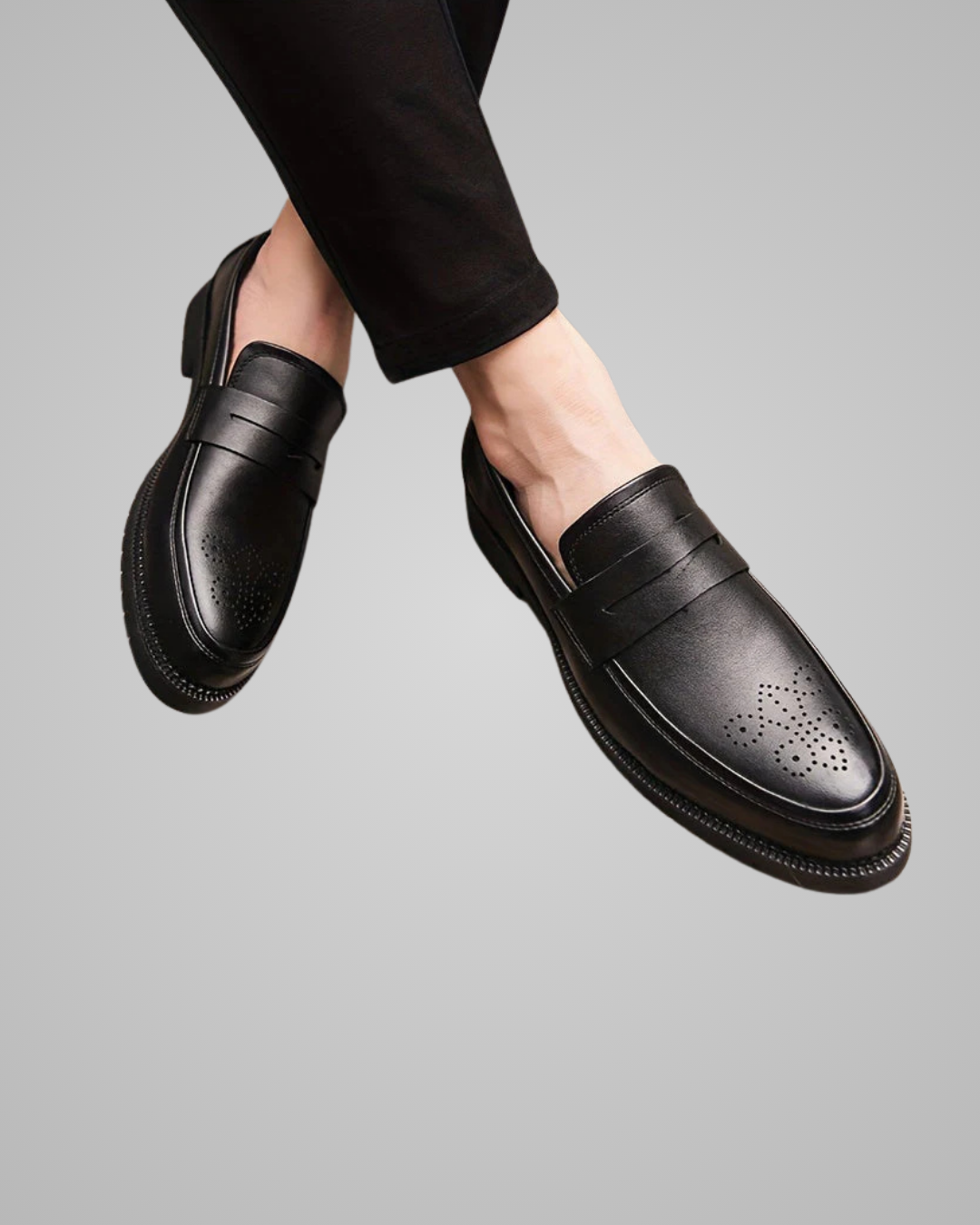 A pair of black leather penny loafers with decorative brogue perforations on the toe, shown on a model's feet against a solid gray background.