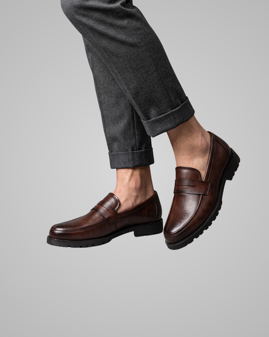 A person wearing dark brown leather penny loafers with dark gray cuffed trousers, shown from the knees down against a solid light gray background.