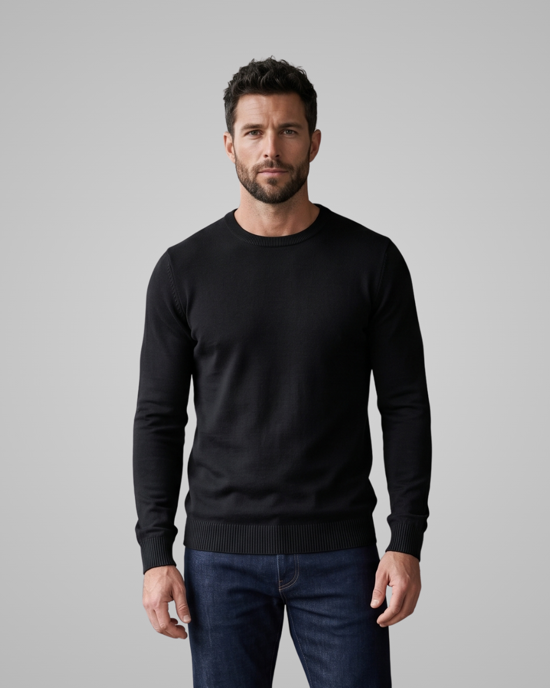 Man wearing a black sweater and blue jeans against a gray background