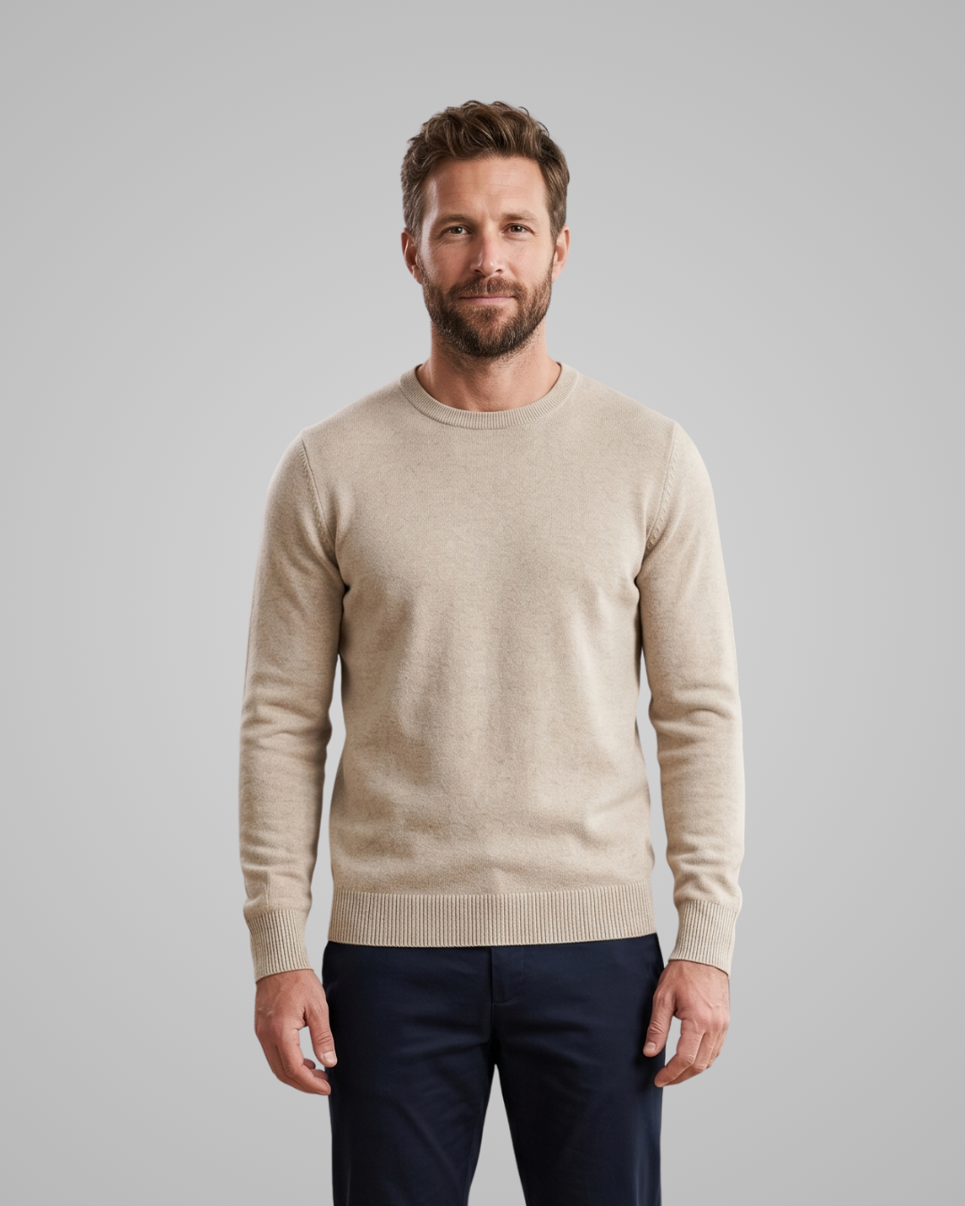 Man wearing a beige sweater and dark pants against a gray background