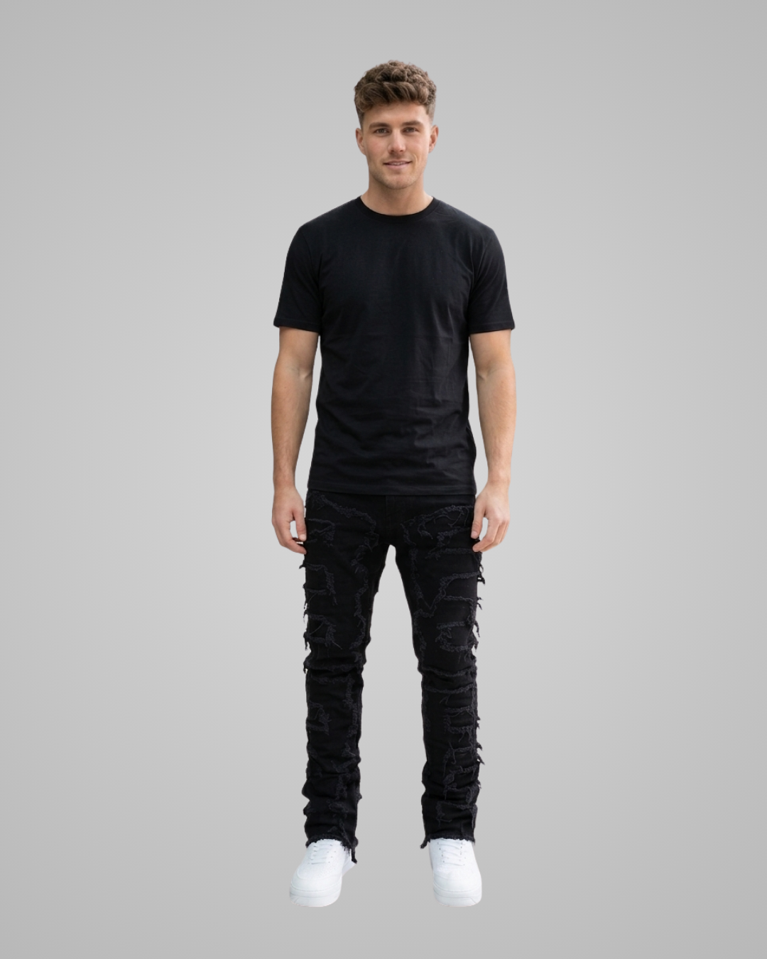 A man wearing a black crew-neck t-shirt, black distressed cargo-style pants with lace-up details, and white sneakers, standing against a solid gray background.