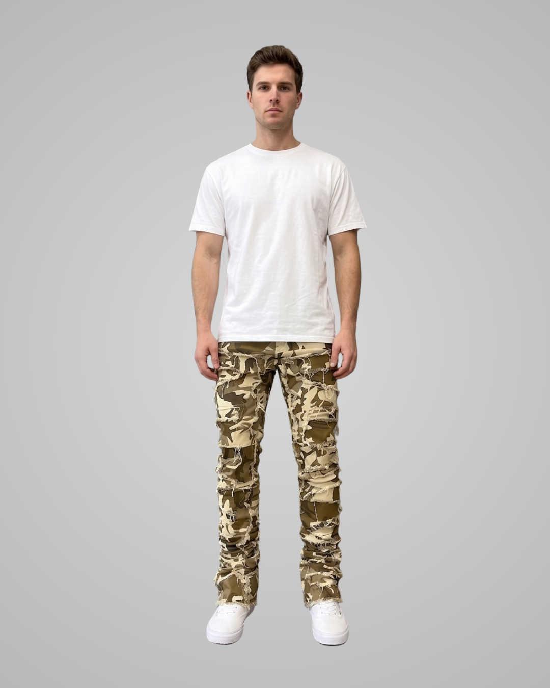 A man stands against a plain light gray studio background wearing a plain white short-sleeve T-shirt, camouflage pants, and white sneakers.