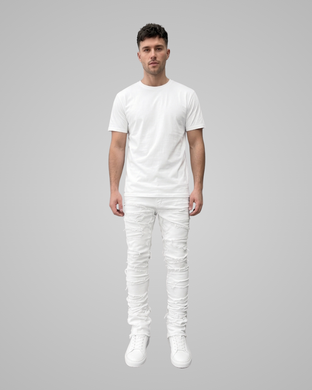 A man modeling a white short-sleeve t-shirt, white distressed skinny jeans, and white sneakers against a solid gray background.