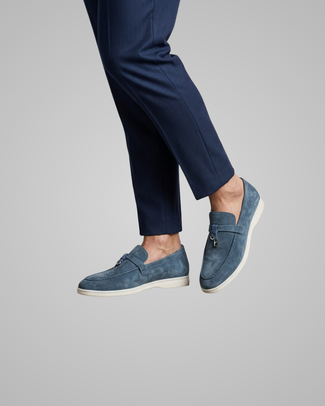 A person wearing navy blue trousers and blue suede slip-on loafers with white soles, shown against a plain light gray studio background.
