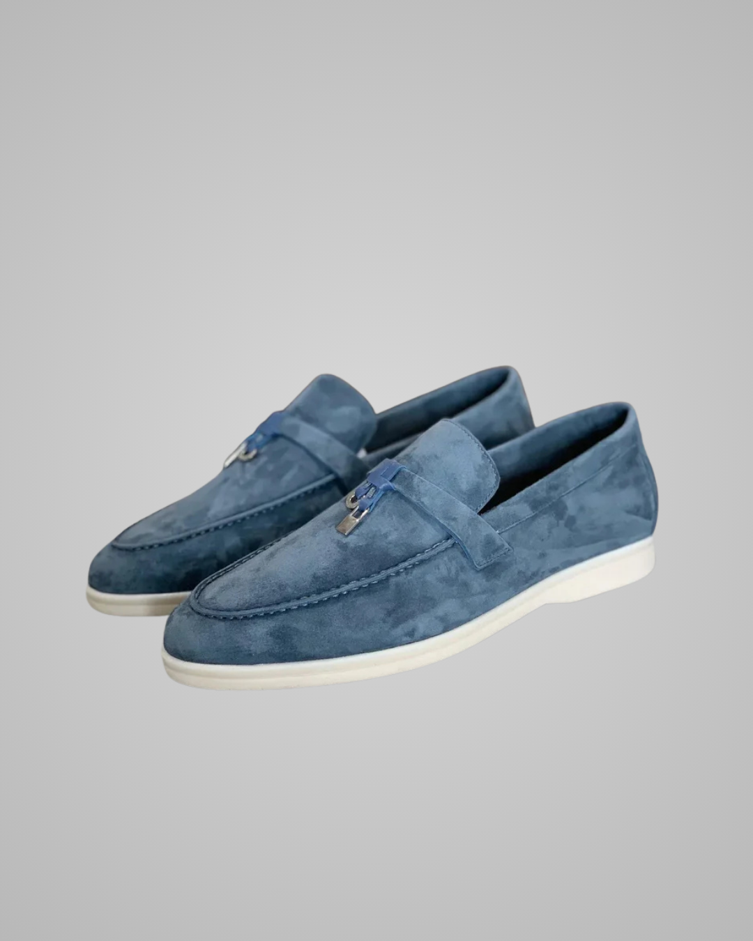 A pair of blue suede slip-on loafers with white soles, displayed against a neutral gray background.