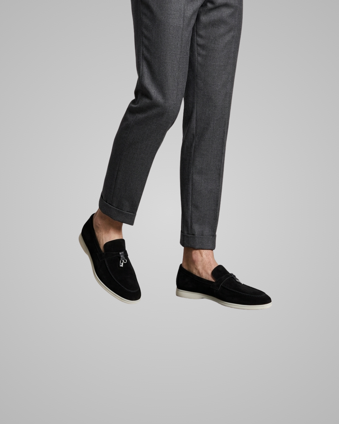 A person wearing slim-fit gray trousers and black suede loafers with white soles, shown from the waist down against a light gray studio background.