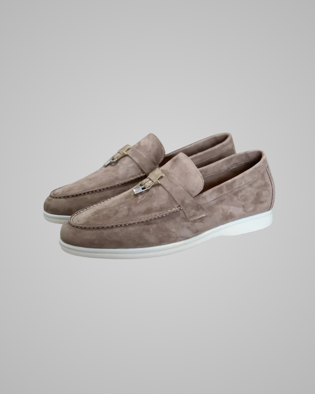 A pair of taupe suede penny loafers with a small gold-tone metal accent on the vamp and a white sole, shown against a neutral gray background.