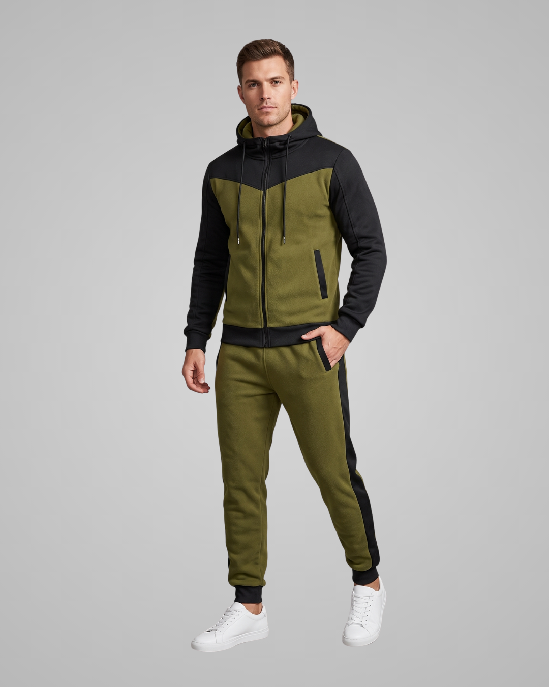 A man modeling a matching olive green and black tracksuit with a zip-up hoodie and jogger pants, paired with white sneakers, against a solid gray studio background.