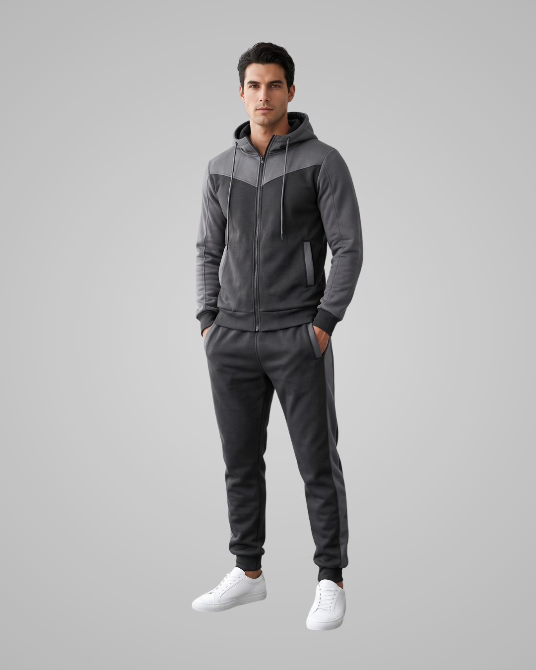 A man modeling a dark gray and black color-blocked zip-up hoodie and matching jogger pants, paired with white sneakers, against a solid gray studio background.