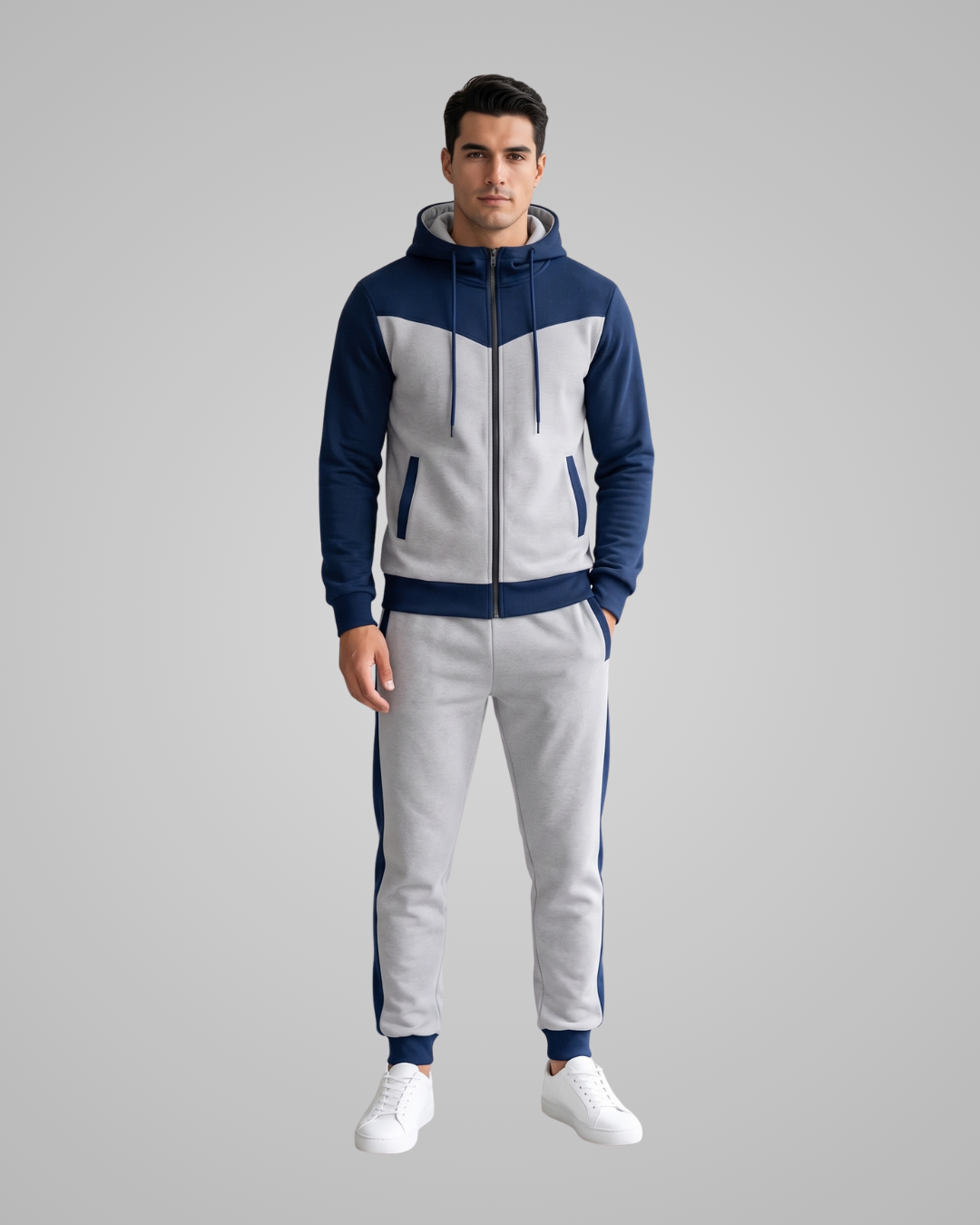 A man modeling a two-piece tracksuit featuring a navy blue and light gray color-blocked zip-up hoodie and matching jogger pants with navy side stripes, set against a solid gray background.
