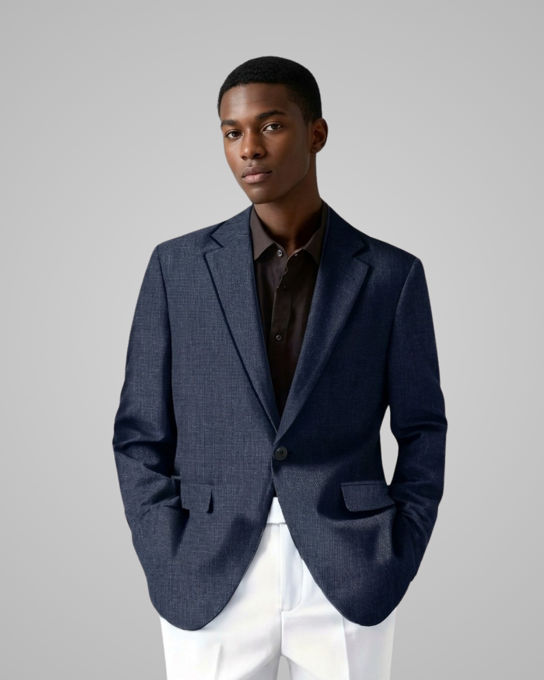 Man wearing a navy blazer over a brown shirt with white pants on a gray background