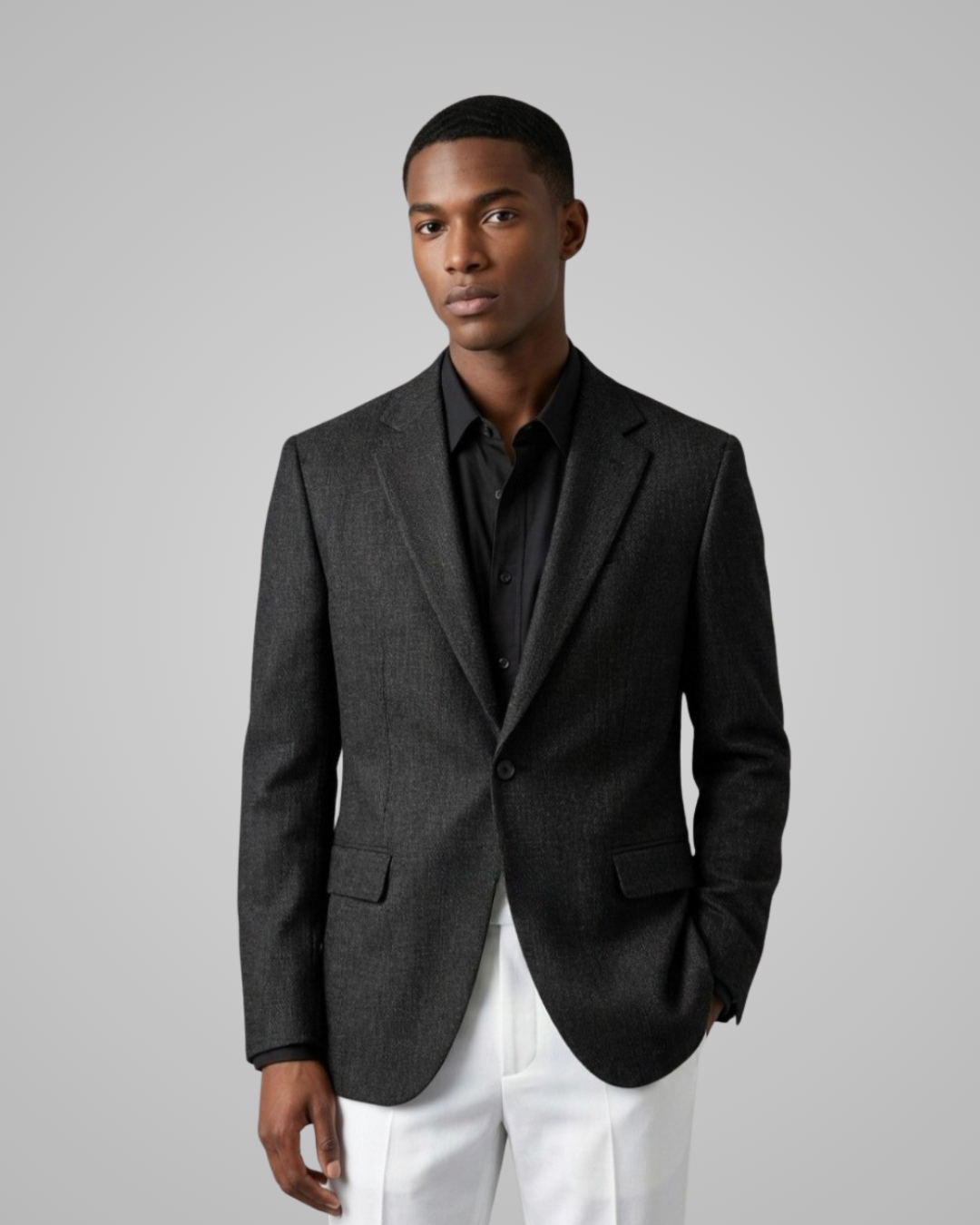 Man wearing a dark gray blazer over a black shirt with white pants on a light gray background