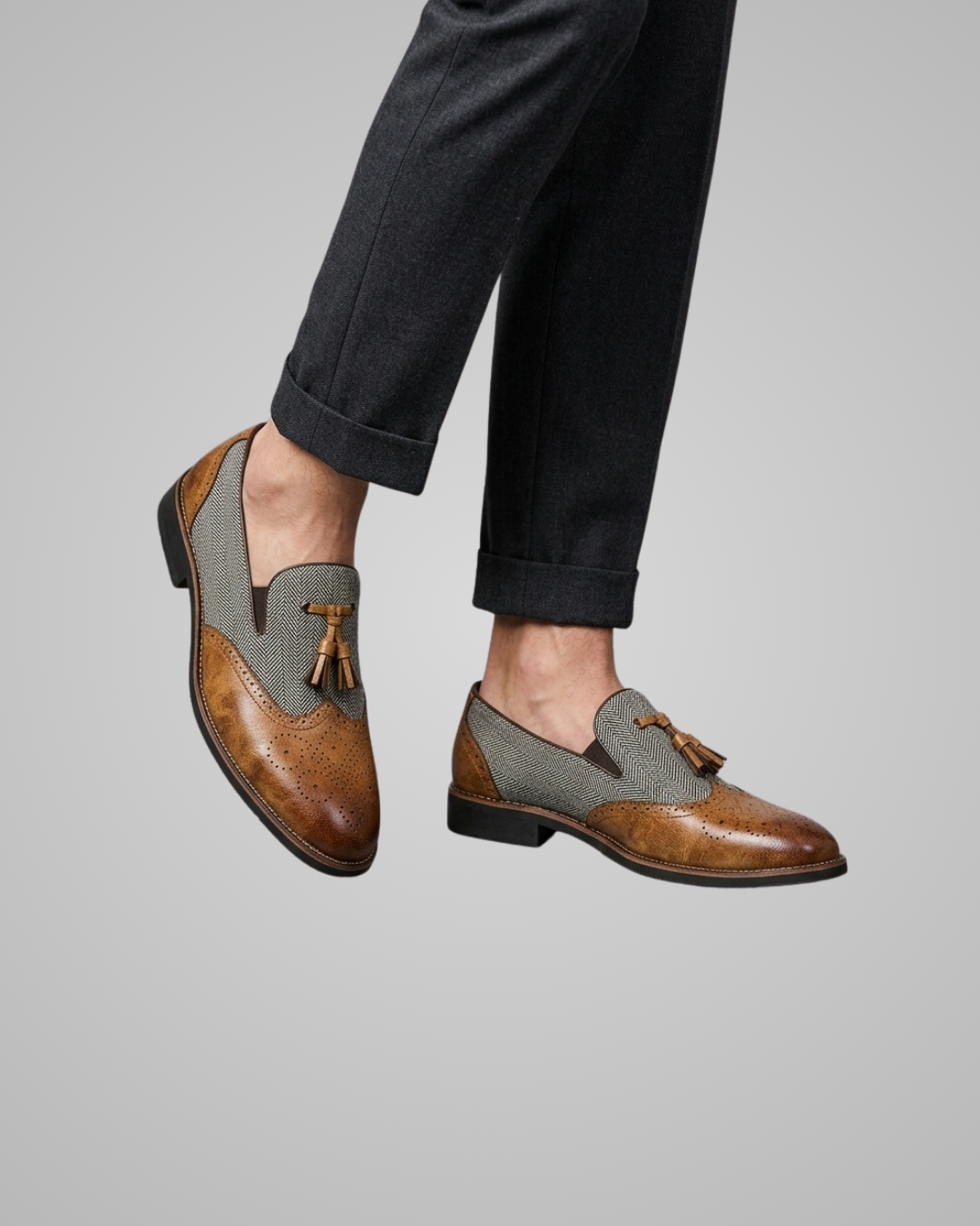 A person wearing brown and gray brogue tassel loafers with dark cuffed trousers, shown against a plain light gray studio background.