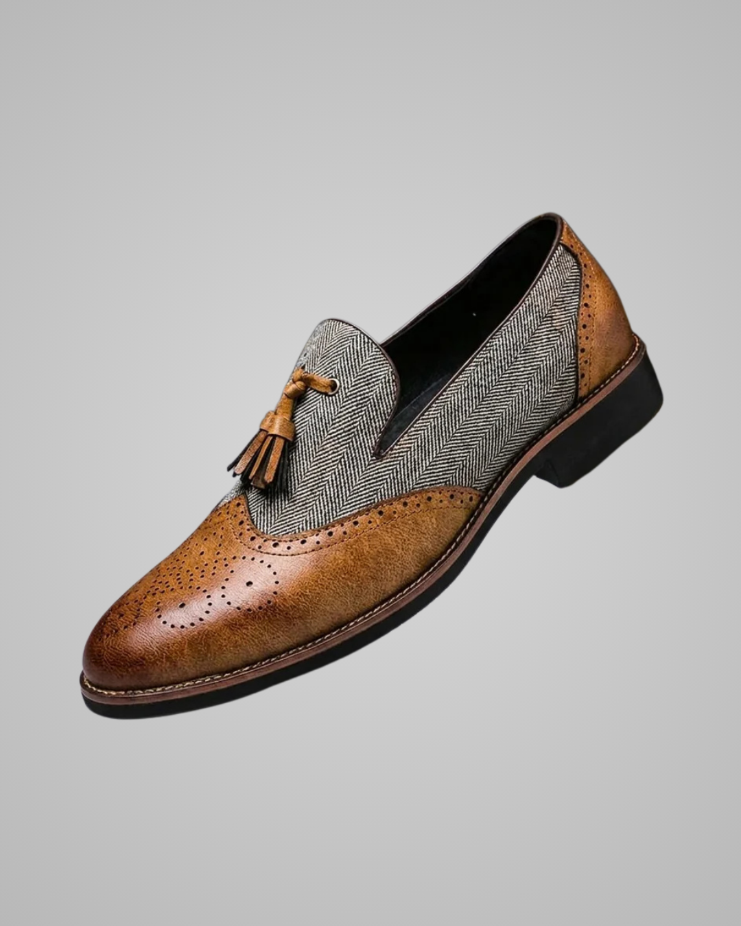 A men's slip-on loafer shoe featuring a brown brogue leather toe and heel, a brown leather tassel detail, and a gray herringbone patterned slipper vamp, shown against a solid gray background.