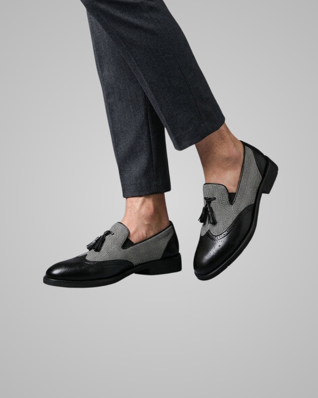 A person wearing dark gray trousers and black-and-gray tassel loafers, shown from the calves down against a plain light gray studio background.
