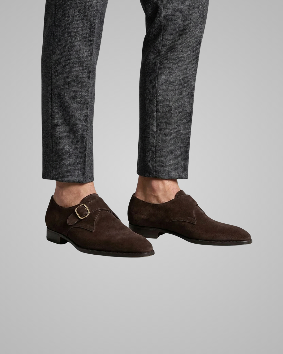 Brown suede shoes with a buckle worn with gray pants on a light gray background