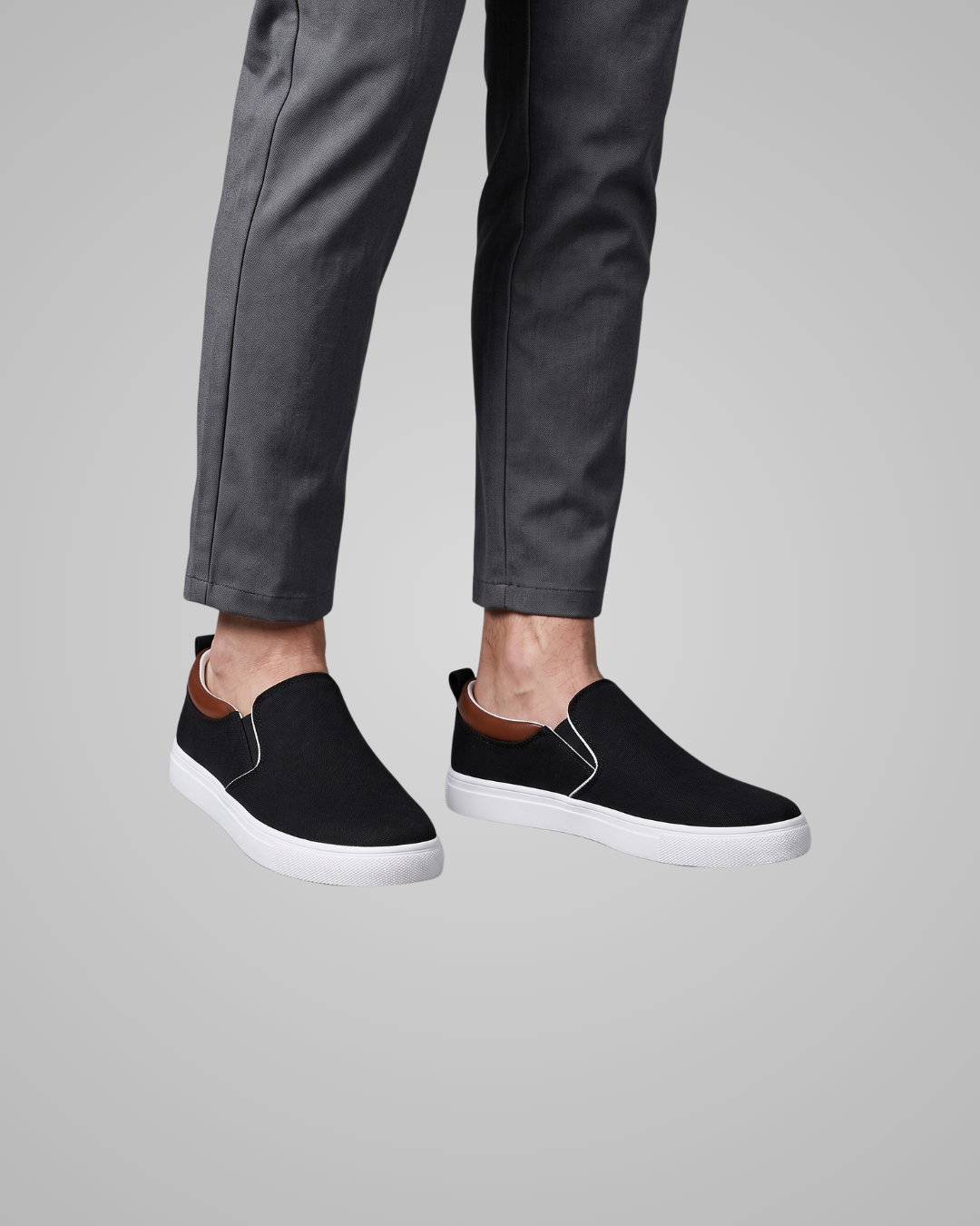 Black slip-on canvas sneakers with white soles and a brown collar lining, shown on a model wearing dark gray trousers against a solid gray background.