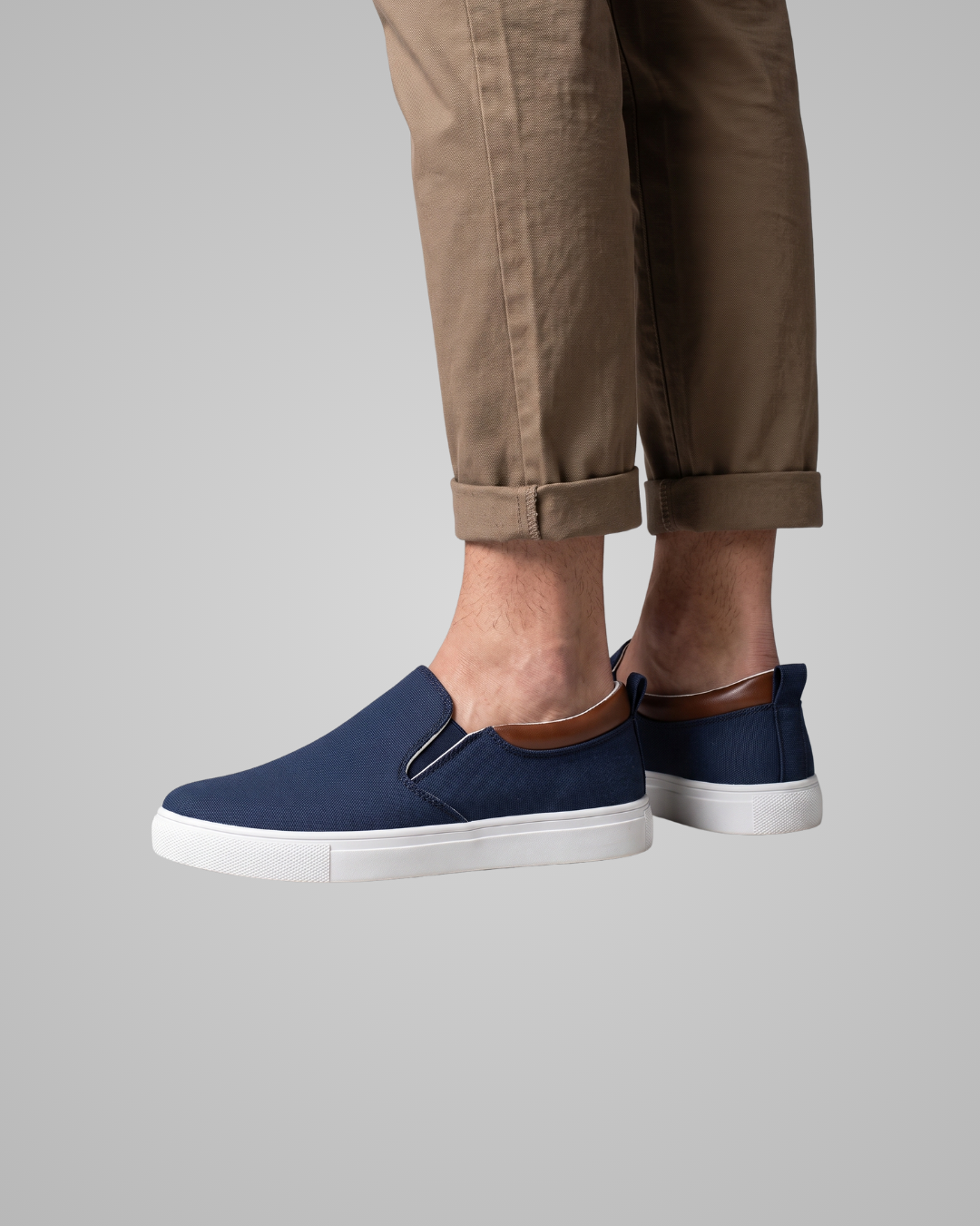 A person wearing navy slip-on sneakers with white soles and brown trim, paired with cuffed khaki pants, shown against a plain gray studio background.