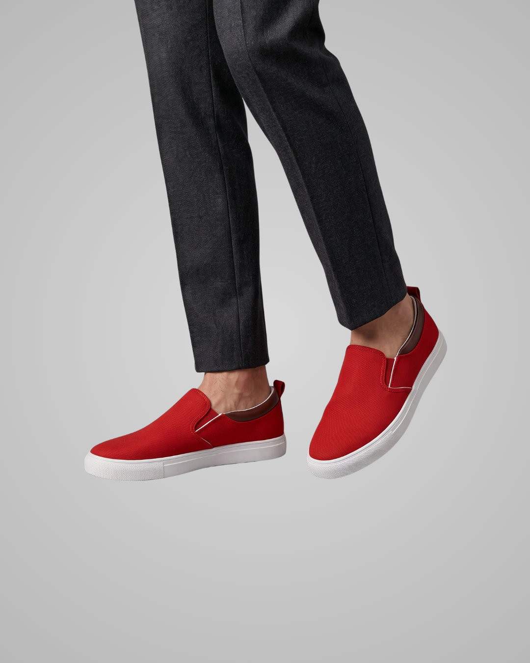 A person wearing dark charcoal trousers and bright red slip-on canvas sneakers with white soles, shown against a plain light gray studio background.