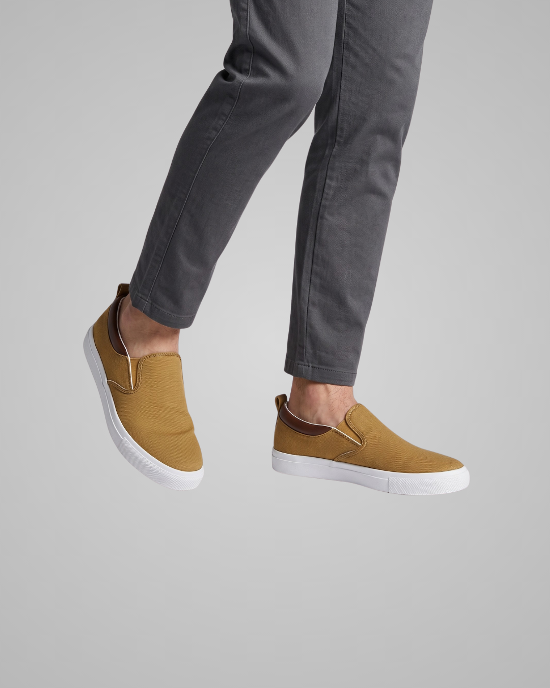 A person wearing slim-fit gray pants and a pair of slip-on canvas sneakers in mustard yellow with white soles and dark brown lining, shown against a plain light gray background.