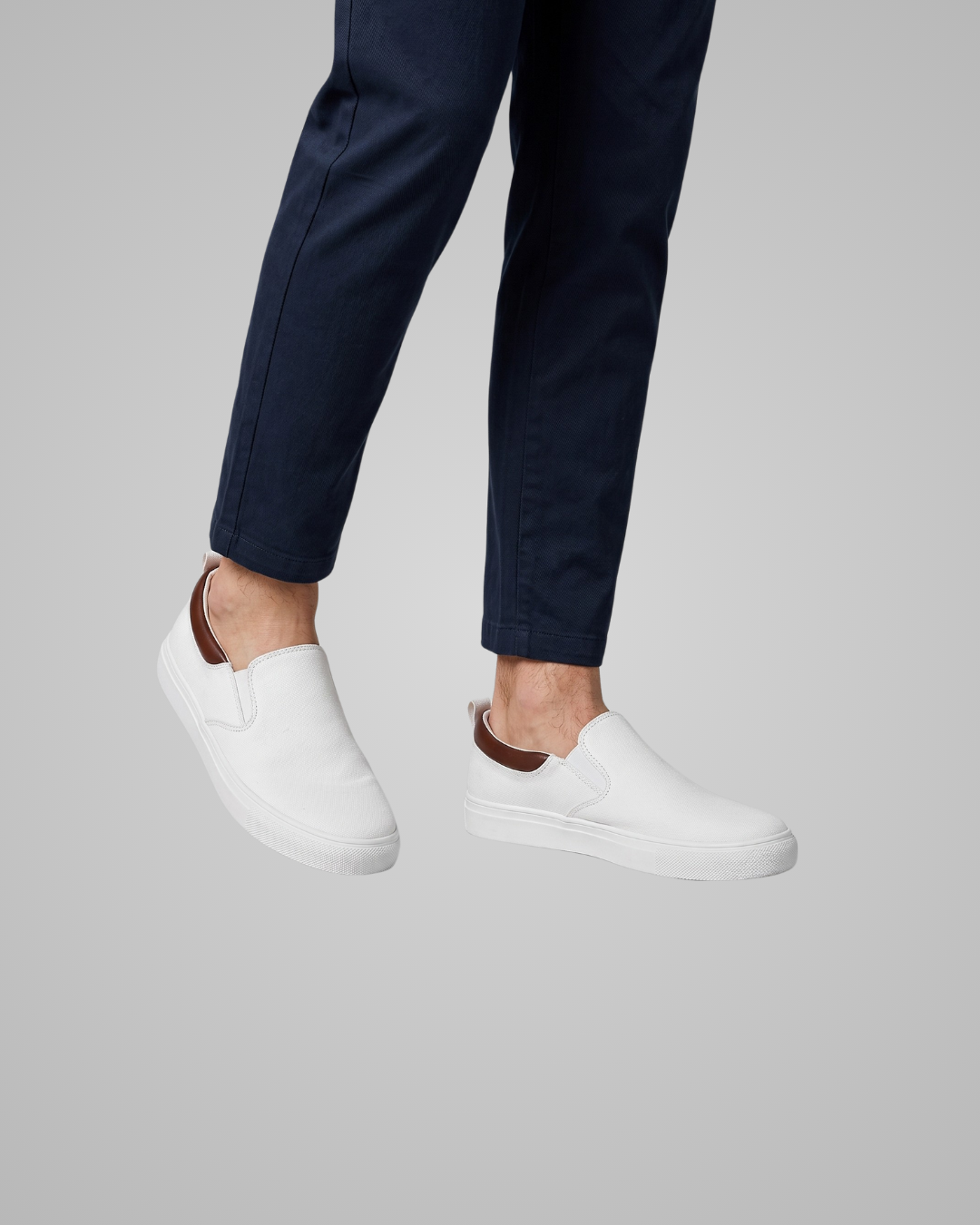 White slip-on sneakers with brown heel accents, shown on a model wearing navy pants against a solid gray background.