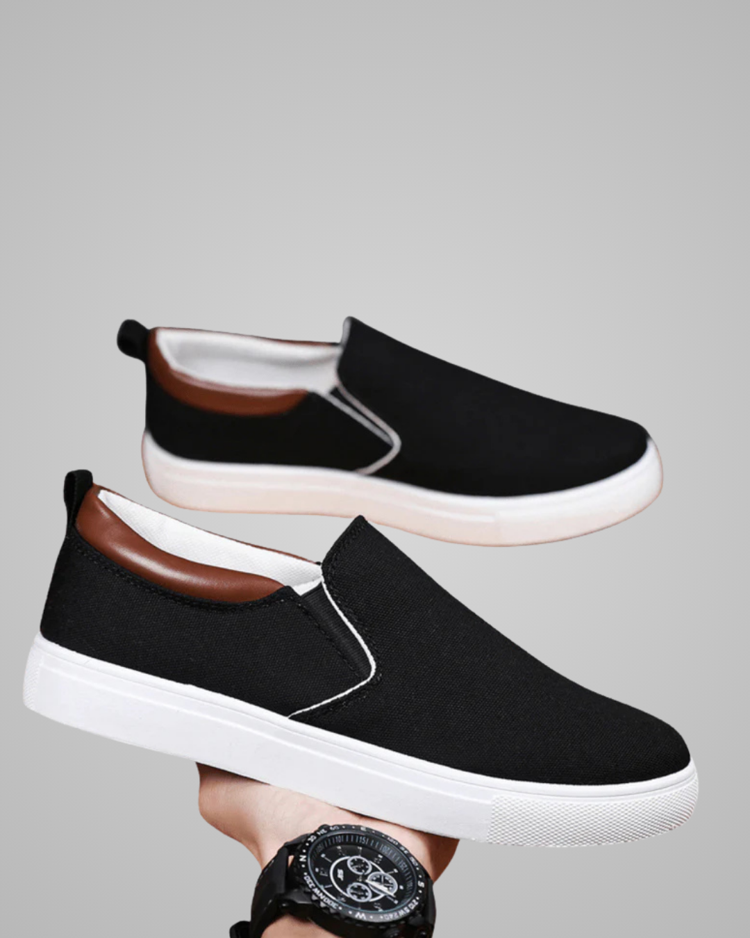 A pair of black slip-on canvas sneakers with white soles and a brown leather-like collar accent, shown against a plain gray background. One shoe is held up by a hand wearing a black watch.