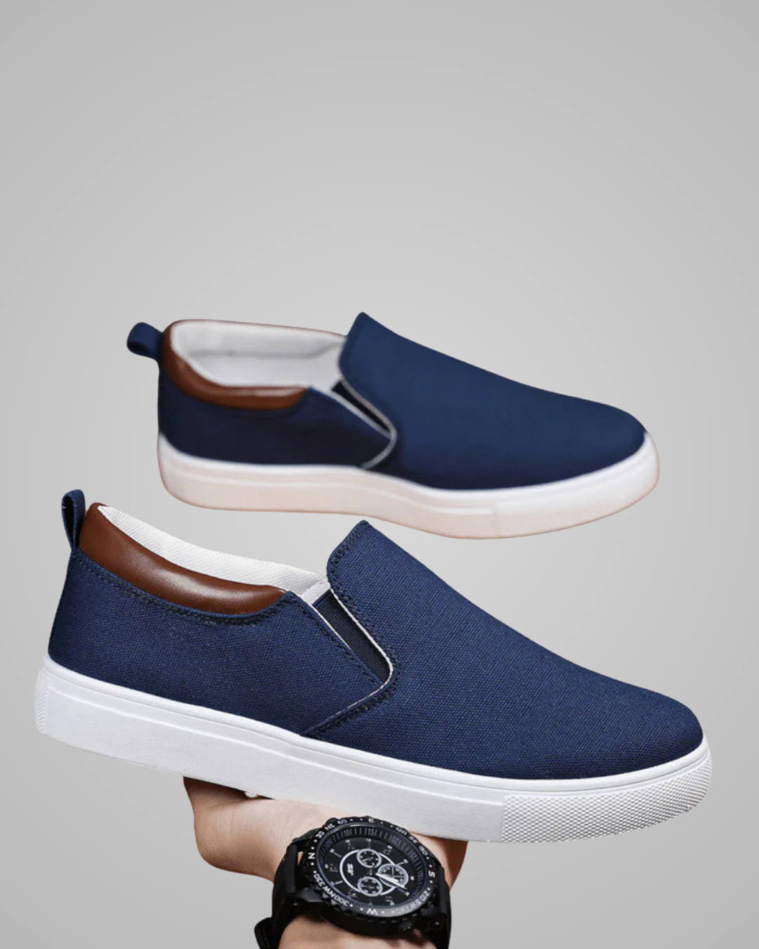 A pair of slip-on canvas sneakers in navy blue with brown heel accents and white soles, shown against a plain gray background. One shoe is floating above, and the other is held by a hand wearing a black watch.